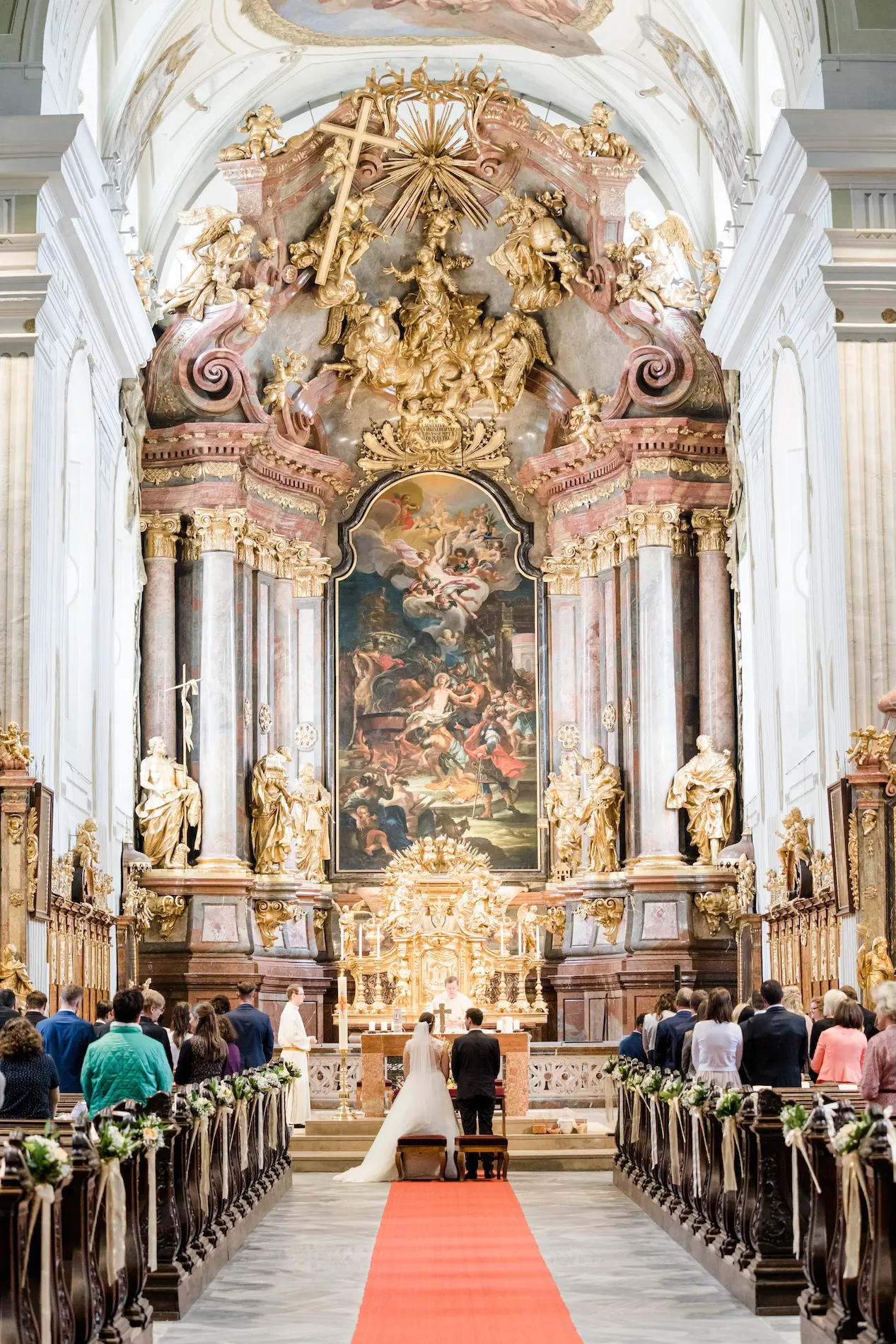 Hochzeitspaar kniet vor einem prachtvollen Altar in einer barocken Kirche, umgeben von Gästen und festlicher Dekoration.