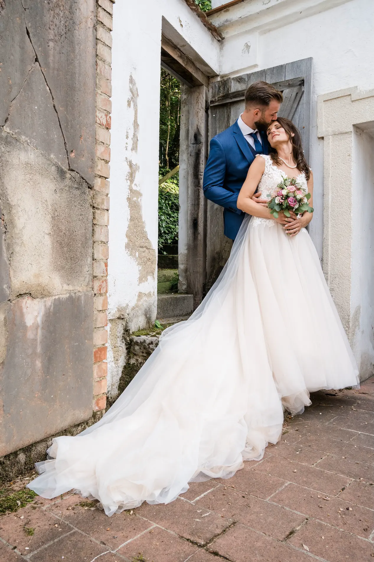 Hochzeitspaar vor einer alten Tür, die Braut in einem weißen Kleid mit langem Schleier hält einen Blumenstrauß, während der Bräutigam sie liebevoll umarmt.