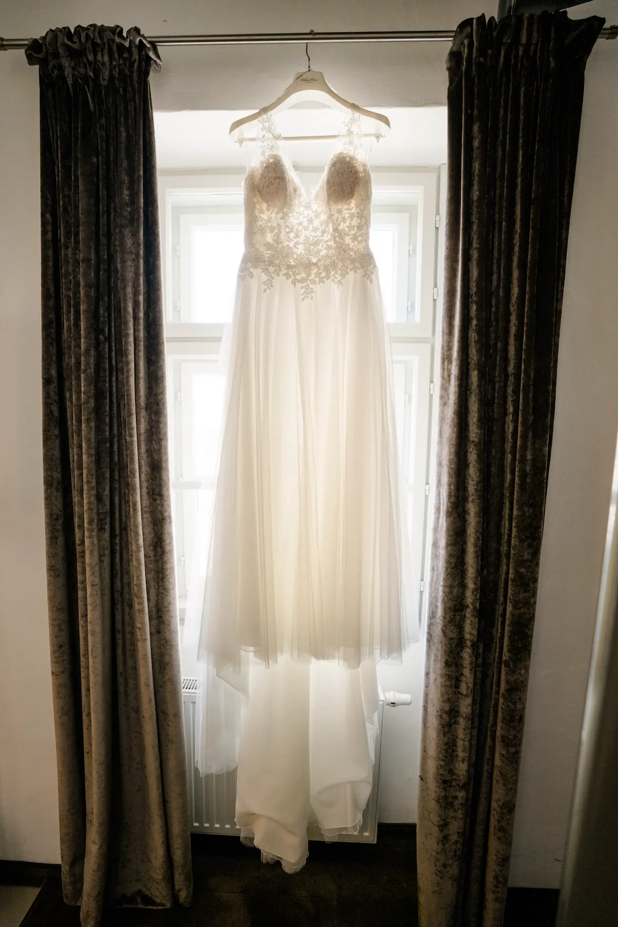 Elegantes Hochzeitskleid aus Spitze und Tüll hängt vor einem hellen Fenster zwischen dunklen Vorhängen.
