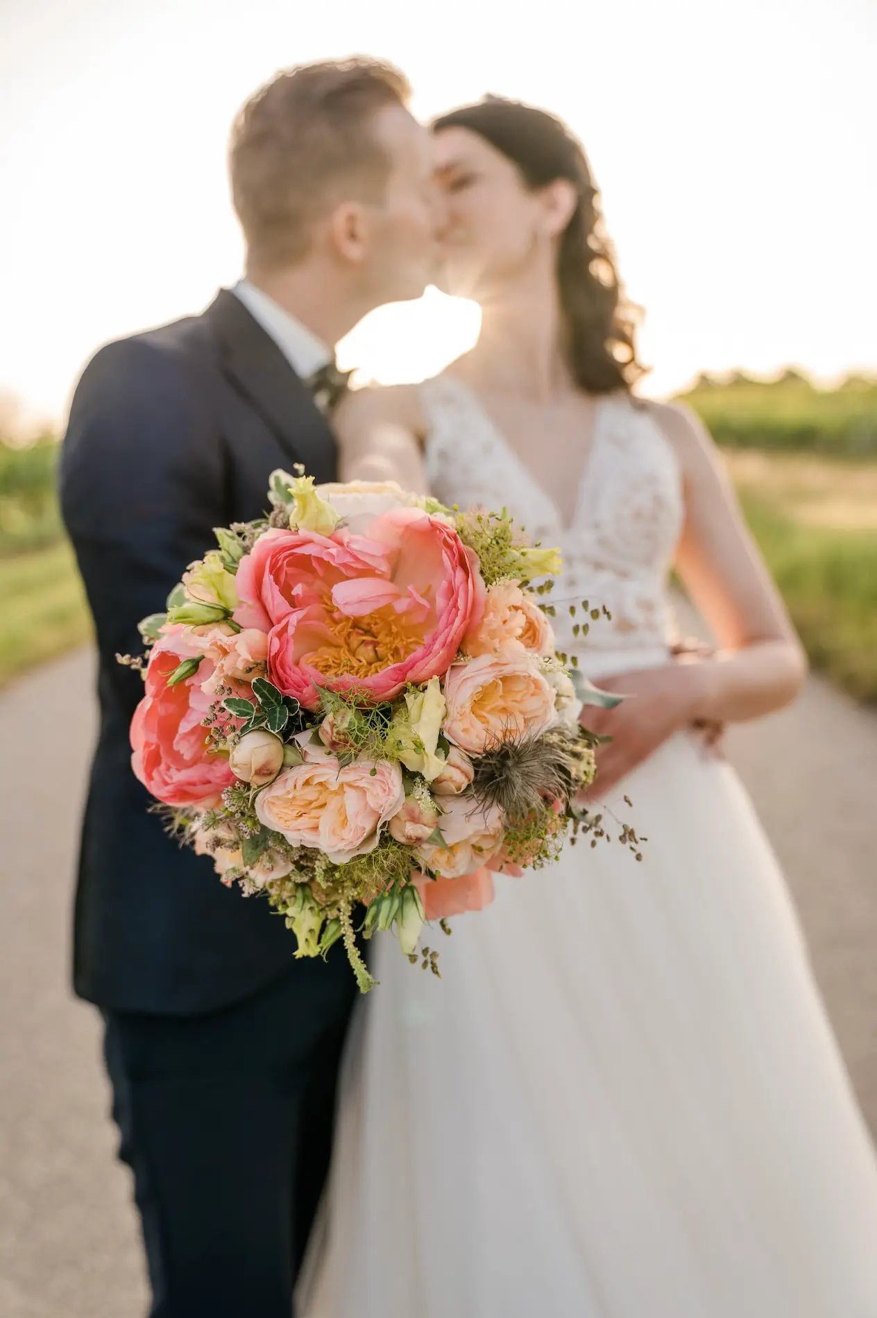 Hochzeitspaar küsst sich bei Sonnenuntergang, mit einem prächtigen Hochzeitsstrauß aus Pfingstrosen und Rosen im Vordergrund.