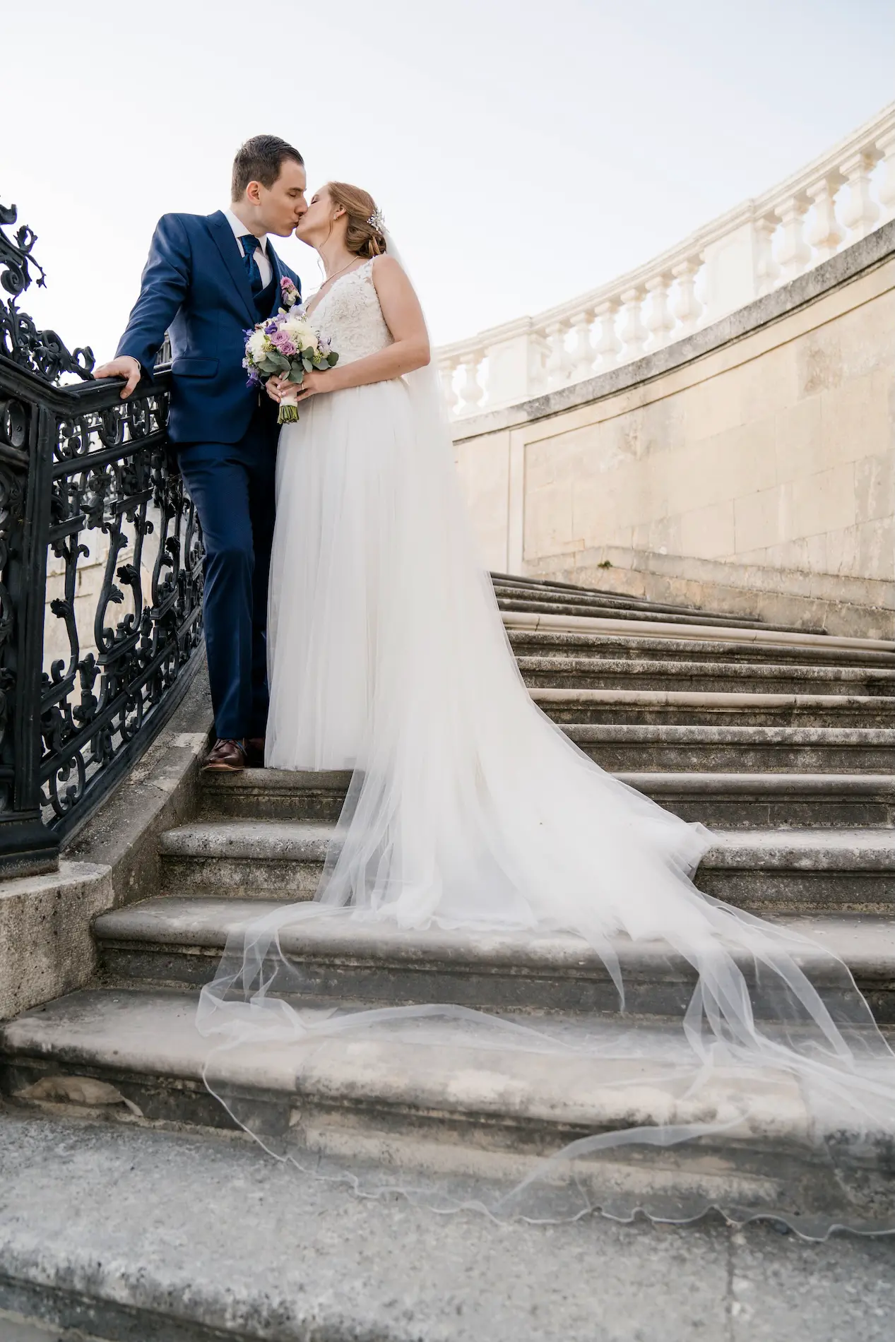 Hochzeitspaar auf einer steinernen Treppe, die Braut im weißen Kleid mit langem Schleier hält einen Blumenstrauß und küsst den Bräutigam im blauen Anzug.