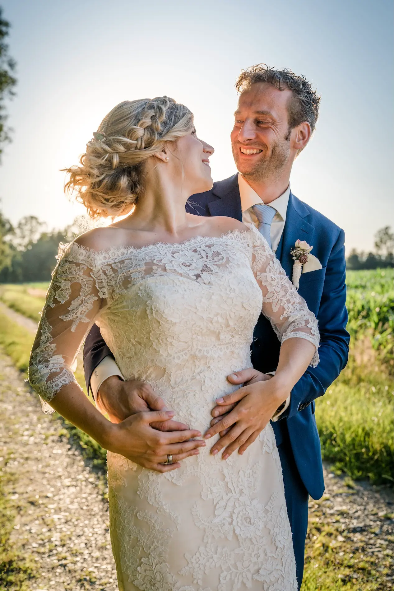 Hochzeitspaar auf einem Weg bei Sonnenuntergang, die Braut im Spitzenkleid wird liebevoll vom Bräutigam umarmt.