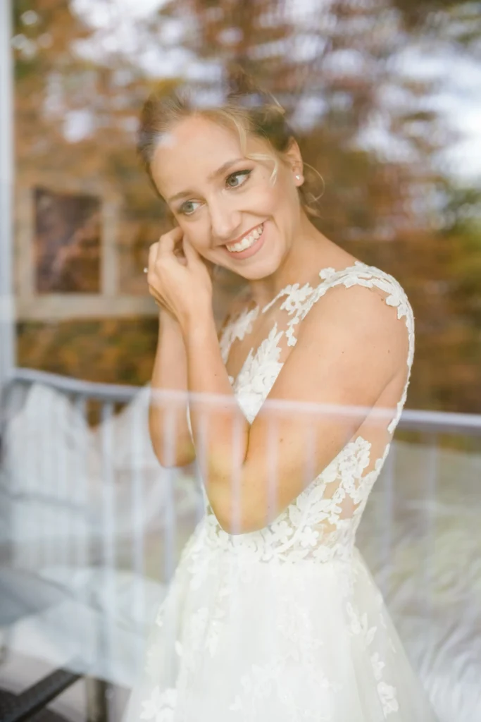 Braut im Spitzenhochzeitskleid, lächelnd beim Einsetzen von Ohrringen, durch ein Fenster fotografiert.