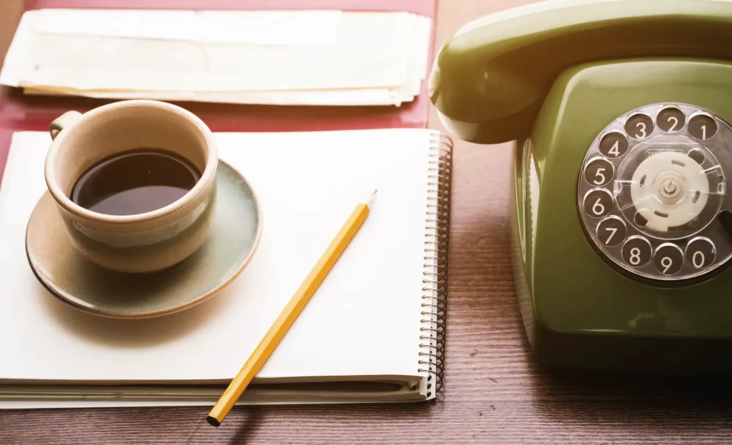 Tasse schwarzer Kaffee, gelber Bleistift, Notizblock und ein grünes Retro-Wählscheibentelefon auf einem Holztisch.