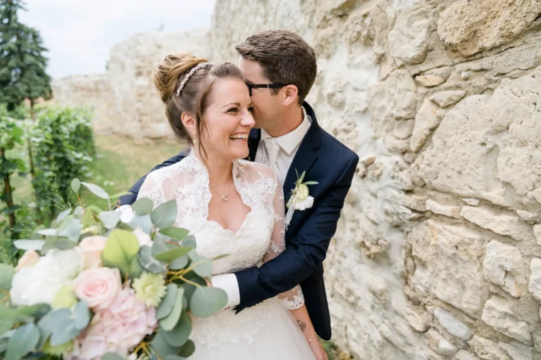 Lachendes Brautpaar in Hochzeitskleidung, stehend vor einer Steinmauer, die Braut hält einen Blumenstrauß mit Rosen und Eukalyptus.
