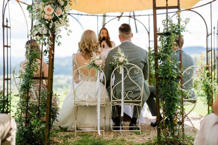 Braut und Bräutigam sitzen während der Trauung unter einem floral geschmückten Pavillon, mit Blick auf eine malerische Landschaft im Hintergrund.